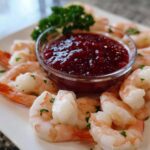 A white platter featuring perfectly cooked shrimp surrounding a glass bowl of deep red shrimp cocktail sauce.