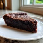 A close-up of a dense, dark slice of flourless chocolate cake resting on a white plate.