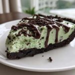 A close-up of a slice of light green grasshopper pie with a dark chocolate crust, topped with chocolate crumbles and fudge drizzle.
