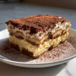 A perfect slice of homemade tiramisu recipe showing layers of cream, ladyfingers, and a heavy dusting of cocoa powder.