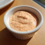 A small white ramekin filled with creamy, orange-hued smash burger sauce speckled with herbs and red pepper flakes.