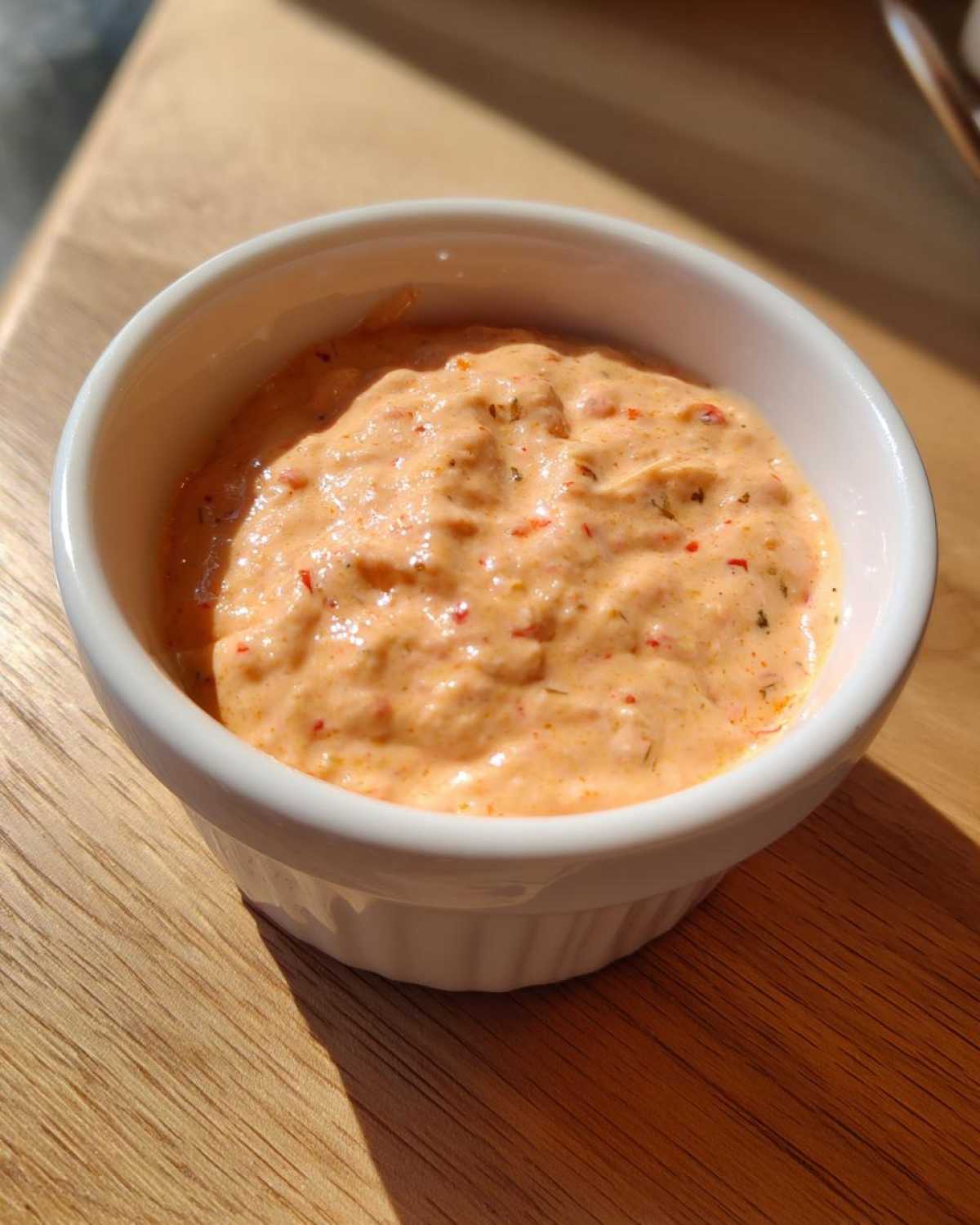 Close-up of creamy, orange-hued smash burger sauce speckled with herbs and spices in a small white bowl.