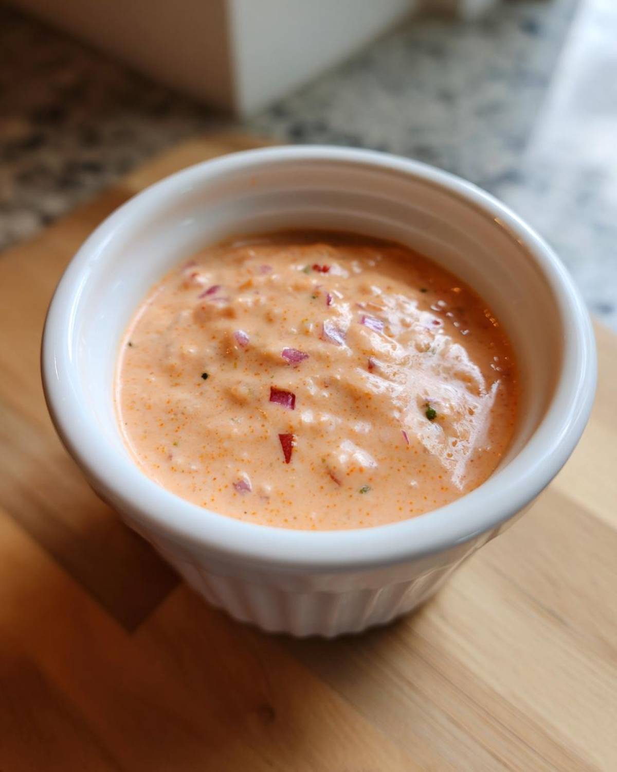 Close-up of creamy, orange-hued smash burger sauce with visible red onion pieces in a white dipping cup.