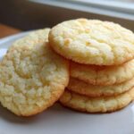 A close-up stack of five golden, soft sugar cookies resting on a white plate near a window.