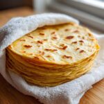 A warm stack of freshly made corn tortillas wrapped in a white cloth on a wooden surface.