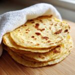 A warm stack of freshly made corn tortillas wrapped partially in a white towel on a wooden surface.
