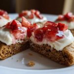 A strawberry cheesecake cookie cut in half showing the soft brown base, cream cheese frosting, and strawberry topping.