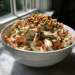 A white bowl filled with creamy, shredded thai chicken salad topped generously with chopped peanuts and green onions.