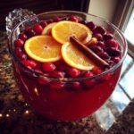 Close-up of a large glass bowl filled with vibrant red Thanksgiving punch, garnished with fresh cranberries, orange slices, and a cinnamon stick.