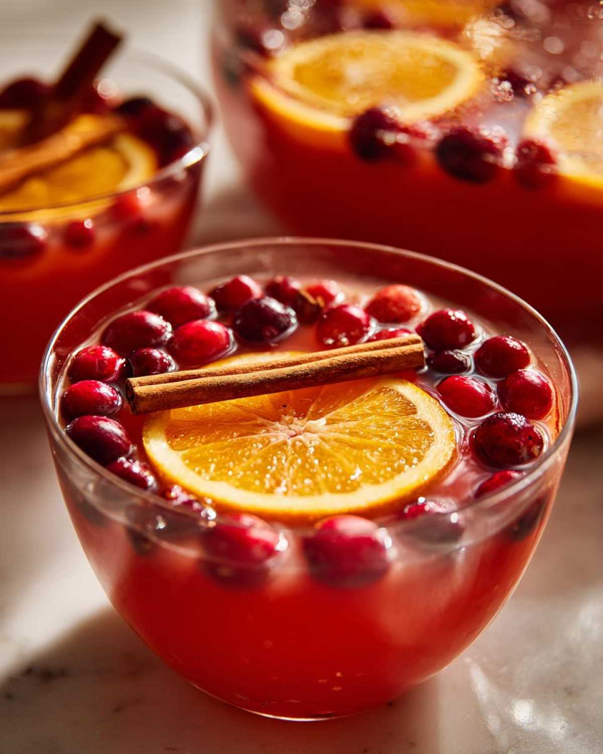 Close-up of a glass filled with bright red Thanksgiving punch, garnished with fresh cranberries, an orange slice, and a cinnamon stick.