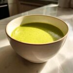 A creamy, bright green bowl of homemade asparagus soup sitting on a sunlit marble counter.