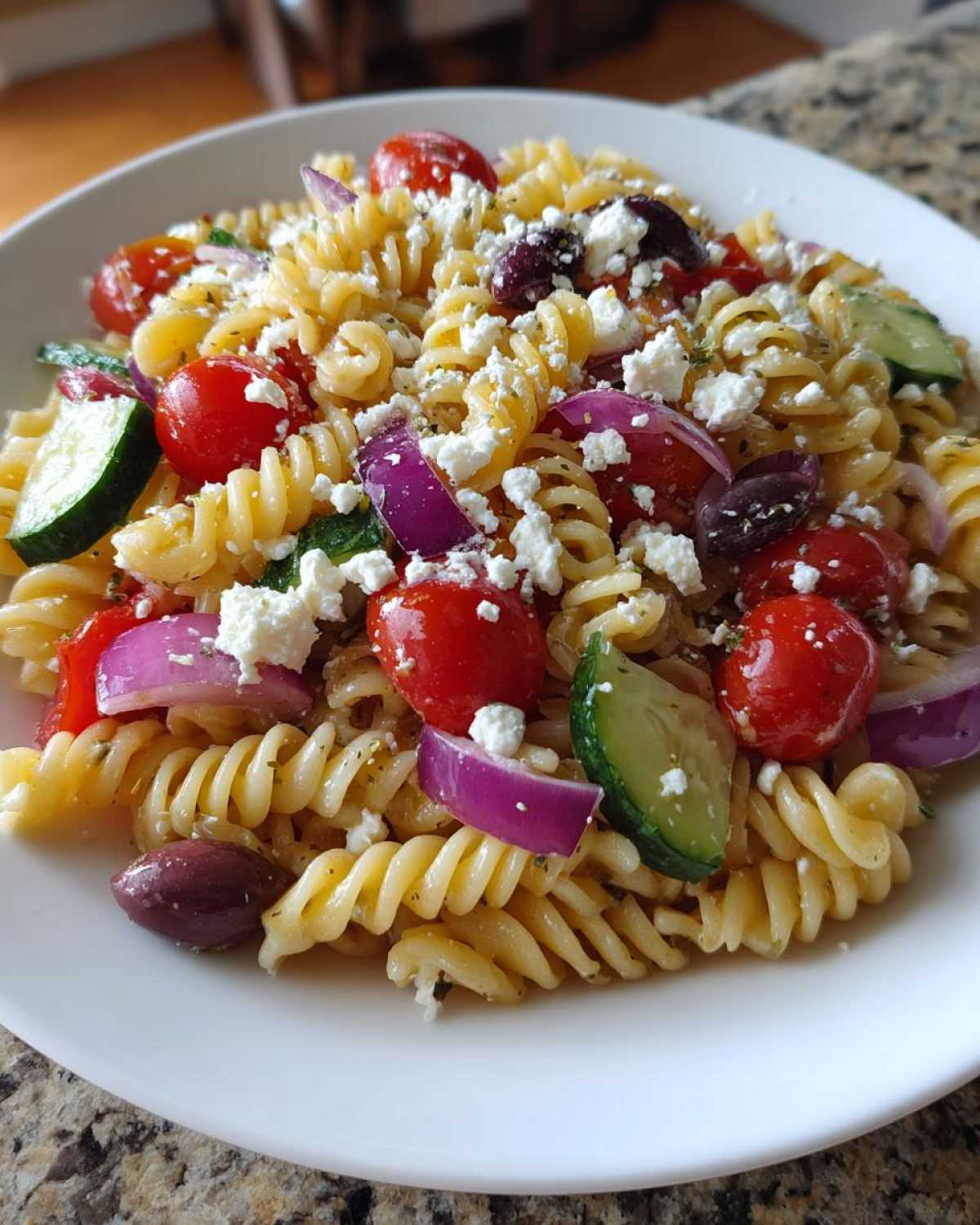 A close-up of a bowl filled with vibrant greek pasta salad featuring fusilli, cherry tomatoes, cucumber, red onion, and feta.