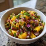 Close-up of a white bowl filled with chunky, colorful peach salsa featuring diced yellow peaches, red onion, and cilantro.