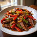 Close-up of glossy pepper steak featuring strips of beef, red and green bell peppers, and onions coated in a rich brown sauce.