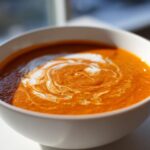 A close-up of vibrant orange roasted tomato soup in a white bowl, swirled with cream.