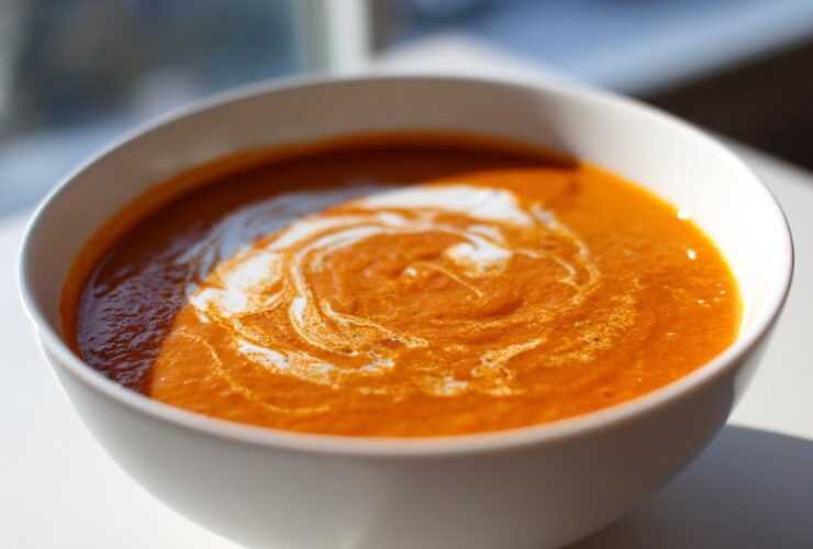 A close-up of vibrant orange roasted tomato soup in a white bowl, swirled with cream.