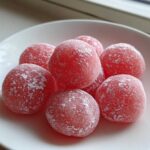 A close-up of several bright pink, round watermelon mochi dusted lightly with white powdered sugar, resting on a white plate.