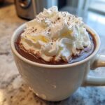 Close-up of a mug filled with rich, dark whipped hot chocolate topped high with fluffy whipped cream and chocolate shavings.