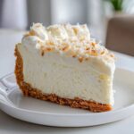 A perfect slice of creamy white chocolate cheesecake with a graham cracker crust, topped with whipped cream and toasted coconut flakes.