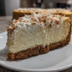 A thick slice of creamy white chocolate cheesecake with a graham cracker crust and topped with whipped cream and toasted coconut.