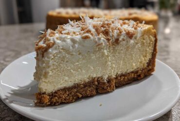 A thick slice of creamy white chocolate cheesecake with a graham cracker crust and topped with whipped cream and toasted coconut.