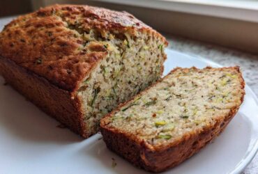 A golden brown loaf of zucchini banana bread sliced open on a white plate, showing moist texture.