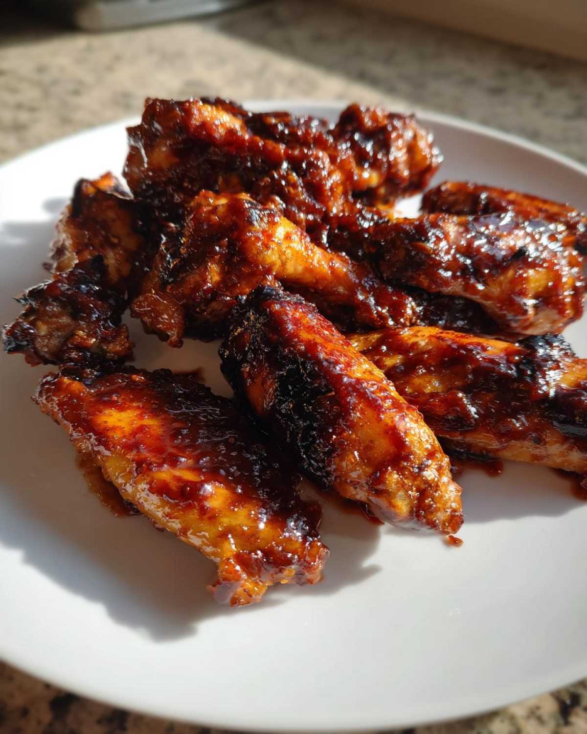 A pile of sticky, dark-glazed Air Fryer BBQ Chicken Wings glistening on a white plate in natural light.