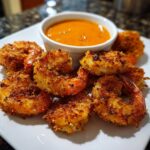 Golden brown Air Fryer Coconut Shrimp arranged on a white plate next to a small bowl of sweet chili dipping sauce.