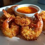 Golden brown, crispy Air Fryer Coconut Shrimp piled on a white plate next to a small bowl of sweet chili sauce.