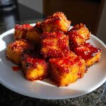 Close-up of golden brown, crispy tofu cubes coated in a shiny, vibrant sweet chili glaze, served on a white plate.