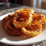 A close-up of golden brown, crispy Air Fryer Onion Rings piled high on a white plate, catching the sunlight.