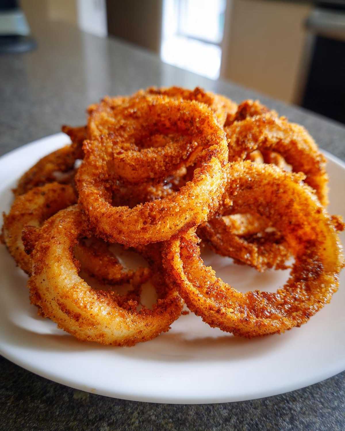 A close-up of a pile of golden brown, crispy Air Fryer Onion Rings seasoned with spices, served on a white plate.
