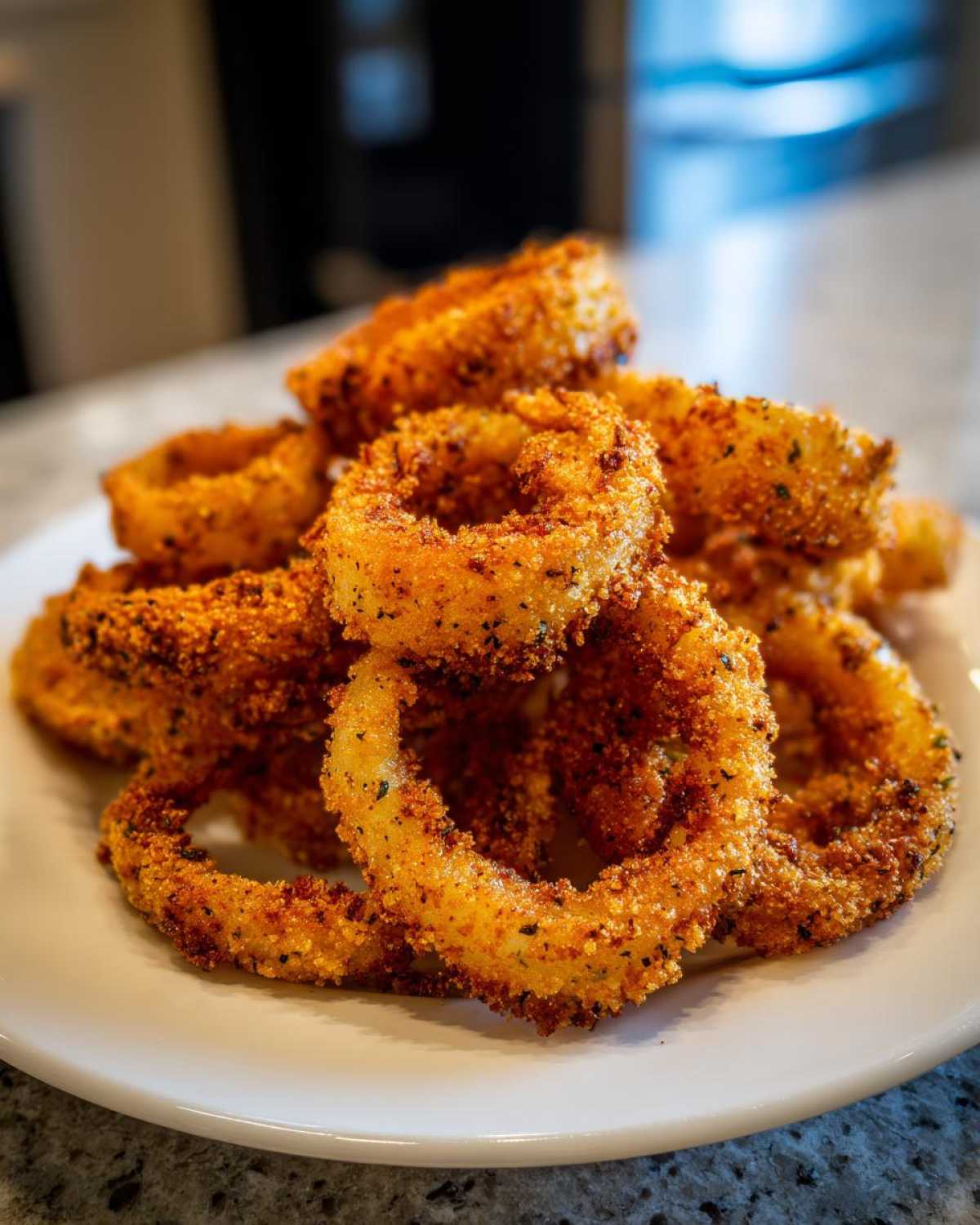 A close-up of a pile of golden brown, crispy Air Fryer Onion Rings seasoned with visible dark specks of herbs and spices.