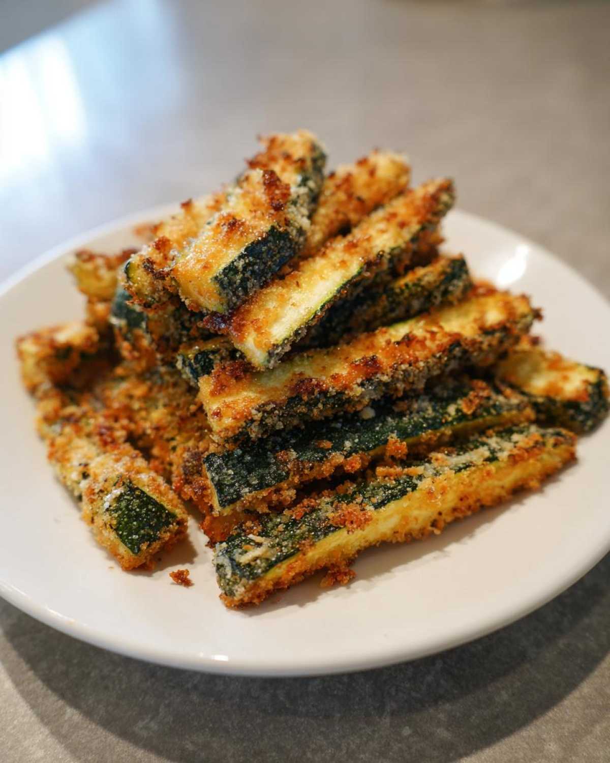 A pile of crispy, golden-brown Air Fryer Zucchini Fries with Parmesan stacked on a white plate.