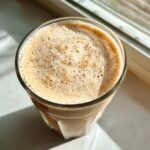 Top-down view of a creamy, frothy Banana Peanut Butter Smoothie in a clear glass catching sunlight.