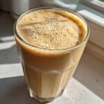 Close-up of a thick, frothy Banana Peanut Butter Smoothie in a tall glass sitting near a bright window.