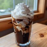A tall glass of rich Iced Mocha swirled with cream and topped high with whipped cream, sitting on a wooden table.