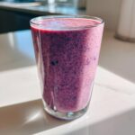A tall glass filled with a thick, vibrant purple Blueberry Lemon Chia Smoothie, sitting on a bright white counter.