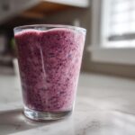 A tall glass filled with a thick, purple Blueberry Lemon Chia Smoothie sitting on a white marble counter.