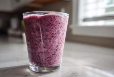 A tall glass filled with a thick, purple Blueberry Lemon Chia Smoothie sitting on a white marble counter.