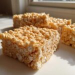 Close-up of several squares of chewy Brown Butter Rice Krispie Treat Bars sitting on a white surface near a window.