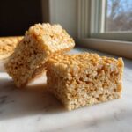 Two thick, chewy Brown Butter Rice Krispie Treat Bars stacked slightly on a white marble surface near a window.