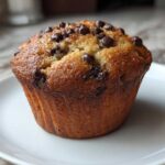 A perfectly baked, golden brown Chocolate Chip Banana Muffin topped with melted chips on a white plate.