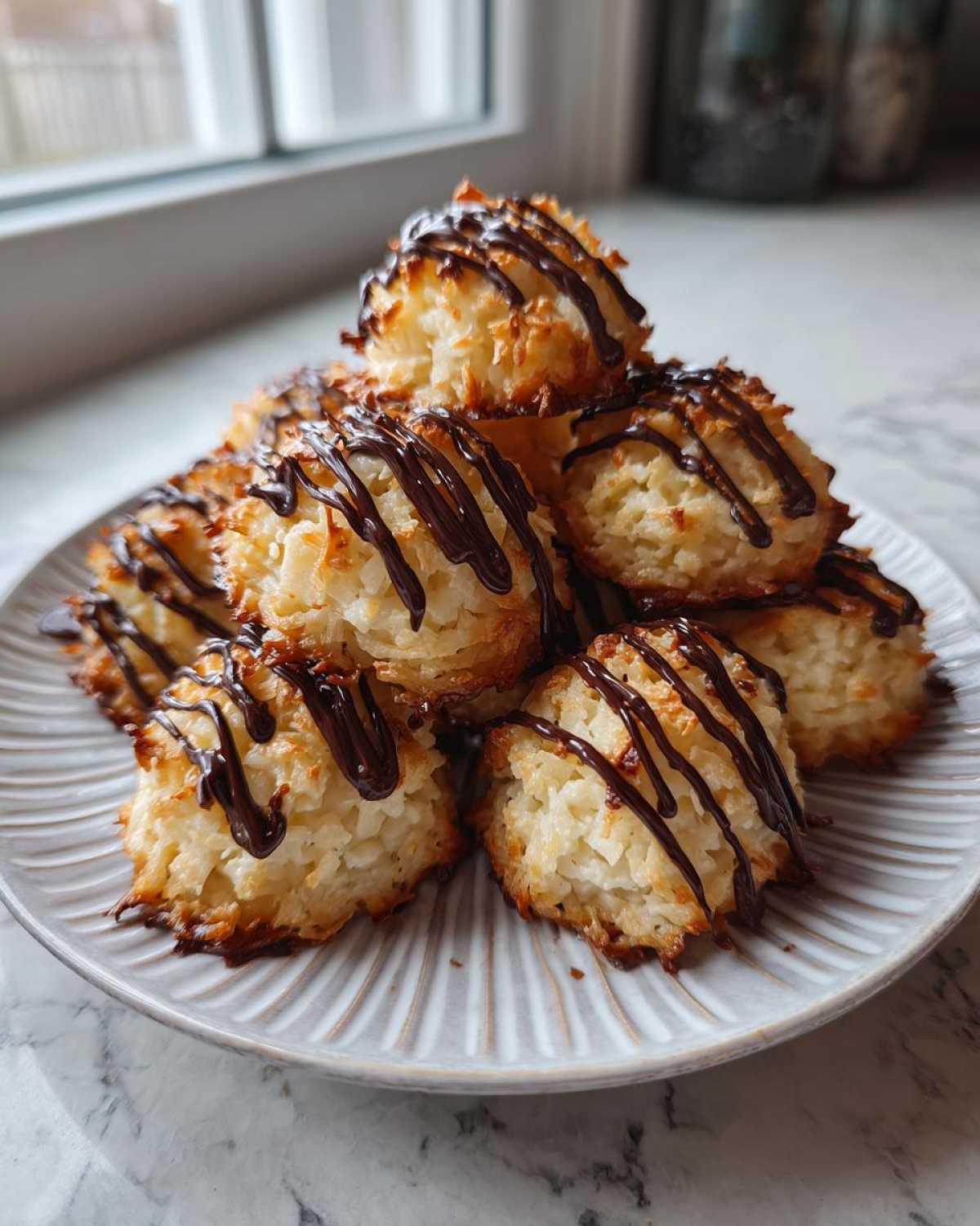 A stack of golden Coconut Macaroons with Dark Chocolate Drizzle piled high on a white, ridged plate.