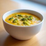 A close-up of a white bowl filled with rich, orange Copycat Broccoli Cheddar Soup topped with bright green broccoli florets.