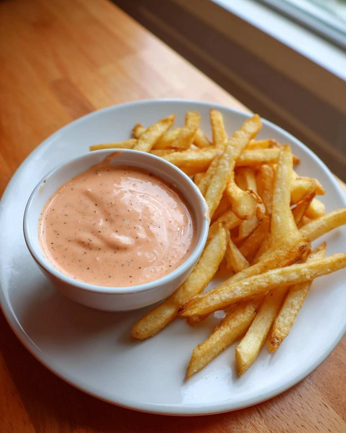 A small white bowl filled with Copycat Raising Cane’s Sauce next to a pile of golden french fries on a white plate.