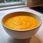 A close-up of a bowl filled with vibrant orange Creamy Coconut Lentil Soup, sitting on a white countertop.