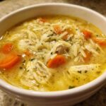 Close-up of a white bowl filled with Creamy Lemon Chicken Orzo Soup, showing shredded chicken, orzo pasta, and bright orange carrots.