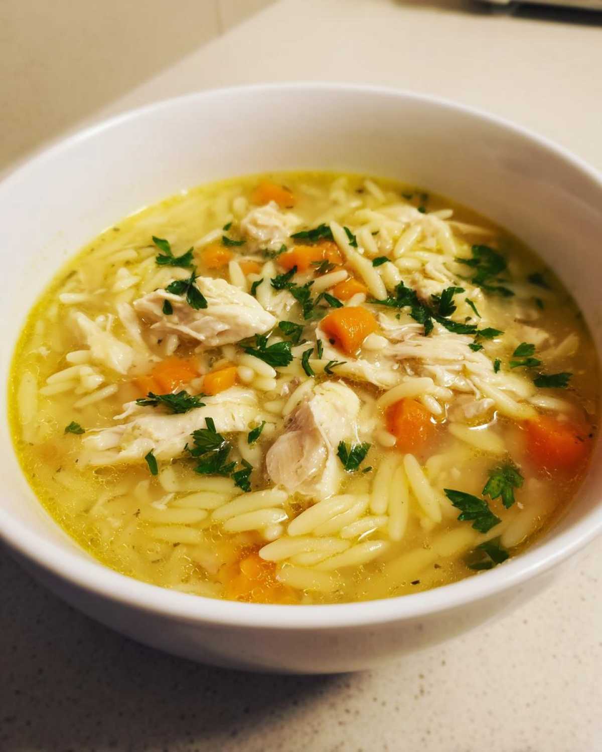 A close-up of a white bowl filled with Creamy Lemon Chicken Orzo Soup, featuring shredded chicken, orzo pasta, carrots, and fresh parsley.