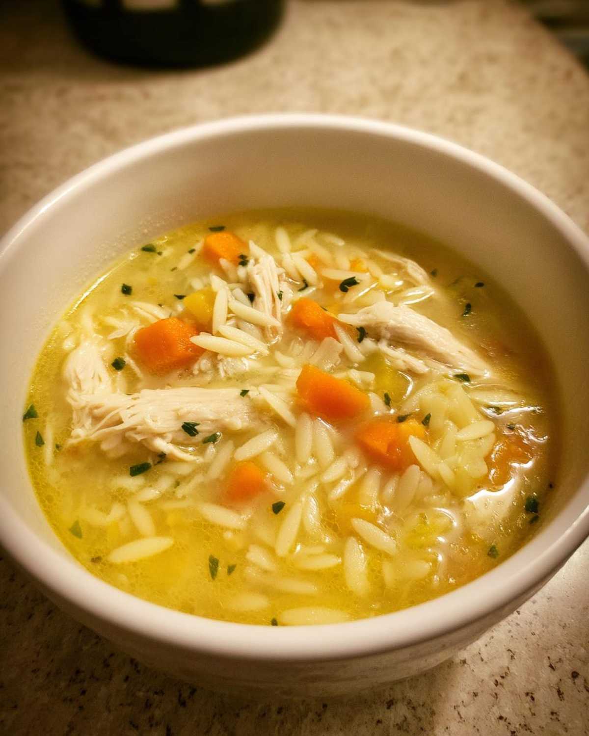 Close-up of a white bowl filled with Creamy Lemon Chicken Orzo Soup, showing shredded chicken, orzo pasta, and diced carrots in a yellow broth.